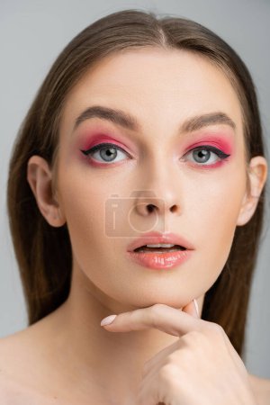 Porträt einer hübschen Frau mit hellem Make-up, die ihr Kinn isoliert auf grau berührt 