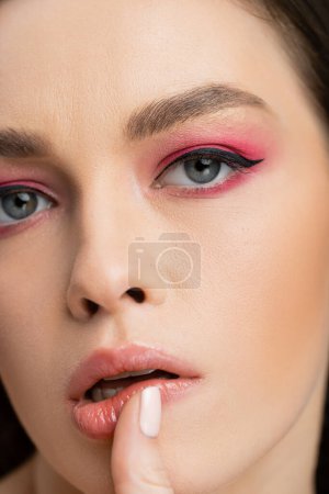 Vista de cerca de una mujer joven con sombra de ojos brillante tocando el labio aislado en gris 