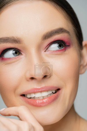 Téléchargez les photos : Close up view of smiling woman with pink eye shadows looking away isolated on grey - en image libre de droit