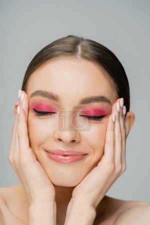Positive woman with bright makeup touching face and closing eyes isolated on grey 