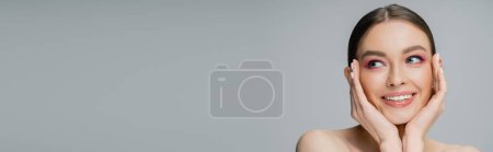 Téléchargez les photos : Jeune femme souriante au visage rose touchant les joues isolées sur gris, bannière - en image libre de droit