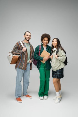 full length of happy and stylish multiethnic students with backpacks and notebook standing on grey background