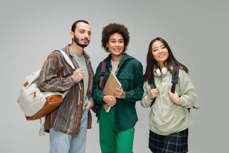 trendy multicultural students with backpacks and notebook smiling and looking away isolated on grey