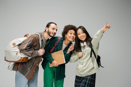 Photo for Cheerful asian woman looking away and pointing with finger near multiethnic students isolated on grey - Royalty Free Image