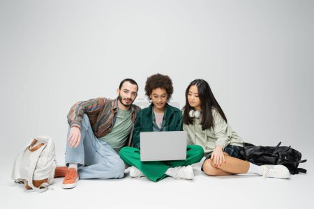 full length of young african american woman sitting with laptop near stylish interracial friends on grey background