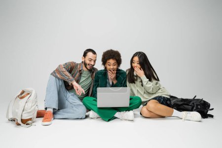 Foto de Longitud completa de amigos multiculturales sorprendidos mirando a la computadora portátil mientras está sentado con las piernas cruzadas sobre fondo gris - Imagen libre de derechos