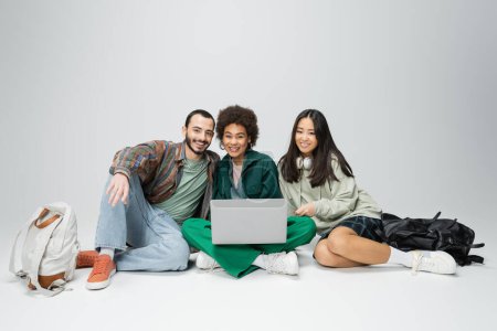 full length of happy and trendy multicultural students sitting with laptop on grey background