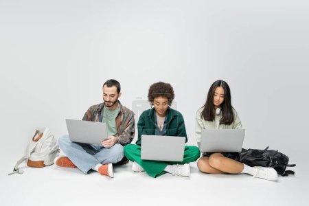 longitud completa de los estudiantes multiculturales que usan computadoras portátiles mientras están sentados con las piernas cruzadas cerca de las mochilas sobre fondo gris