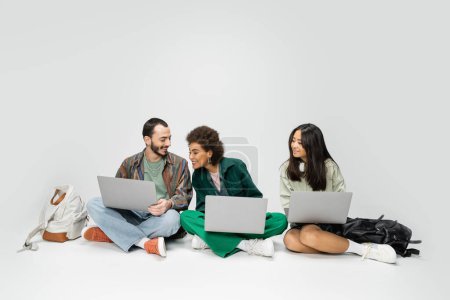 Foto de Longitud completa de estudiantes multiculturales sonrientes sentados con computadoras portátiles cerca de mochilas sobre fondo gris - Imagen libre de derechos