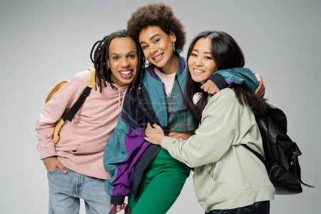 Foto de Happy multicultural friends with backpacks wearing trendy clothes and smiling at camera isolated on grey - Imagen libre de derechos