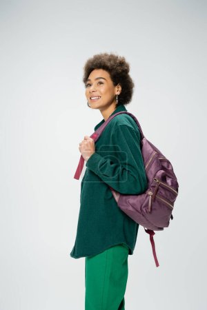 happy and stylish african american woman with backpack looking away isolated on grey
