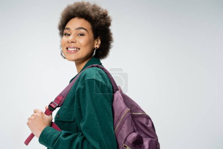 cheerful african american woman in hoop earrings posing with backpack isolated on grey