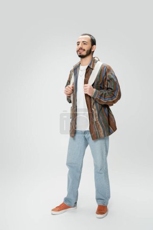 Téléchargez les photos : Homme souriant en jeans bleus et chemise colorée debout avec sac à dos sur fond gris - en image libre de droit