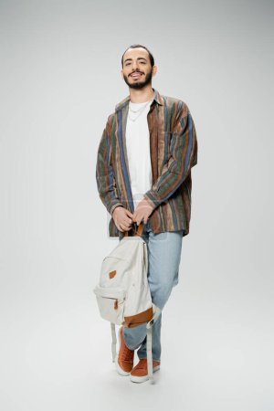 Photo for Full length of cheerful bearded student in trendy outfit standing with backpack on grey background - Royalty Free Image