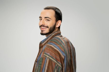 portrait of joyful bearded man in trendy shirt looking at camera isolated on grey