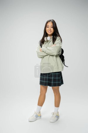 full length of pretty asian student in checkered skirt posing with crossed arms on grey background