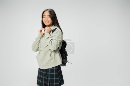 pleased asian woman in hoodie and plaid skirt posing with wireless headphones and backpack isolated on grey