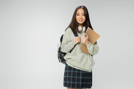 hübsche asiatische Studentin in Kapuzenpulli und kariertem Rock, stehend mit Copybook und Rucksack isoliert auf grau
