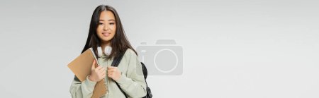 smiling asian woman with wireless headphones holding backpack and notebook isolated on grey, banner