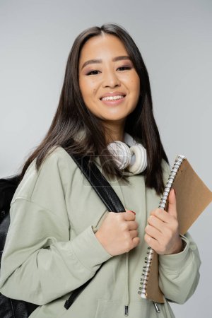 cheerful asian student with wireless headphones and copybook standing with backpack isolated on grey