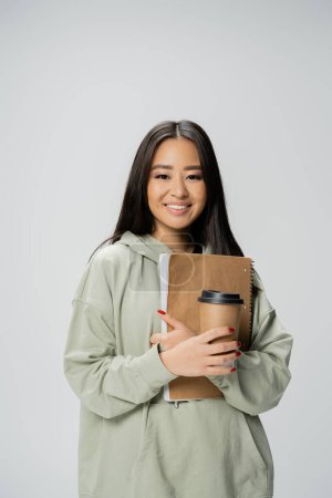 young asian student in hoodie holding takeaway drink and copybook isolated on grey
