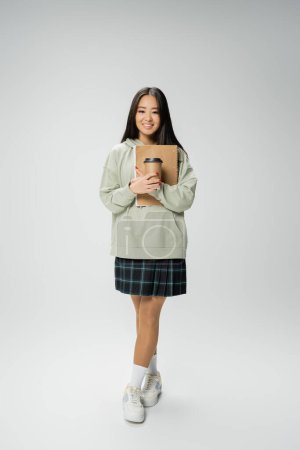 full length of cheerful asian student in checkered skirt and hoodie holding notebook and paper cup on grey background