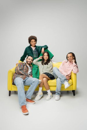 Photo for Fashionable multiethnic friends posing on yellow couch and smiling at camera on grey background - Royalty Free Image