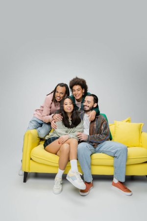 Photo for Smiling multiracial friends in fashionable clothes posing near yellow sofa on grey background - Royalty Free Image