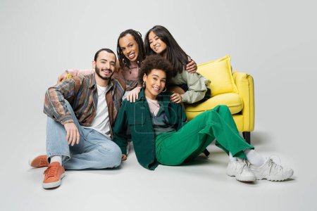 Téléchargez les photos : Amis multiethniques joyeux et élégants regardant la caméra près du canapé jaune sur fond gris - en image libre de droit
