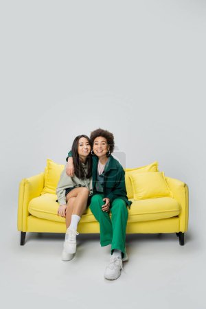 volle Länge der zufrieden afrikanisch-amerikanische Frau schaut in die Kamera und umarmt asiatische Freundin auf gelbem Sofa auf grauem Hintergrund