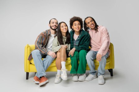 Photo for Full length of smiling multicultural friends in trendy clothes watching movie on yellow sofa on grey background - Royalty Free Image