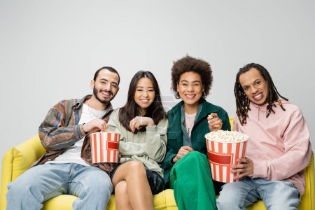 glückliche und stilvolle multiethnische Freunde sitzen auf einer gelben Couch mit Eimern Popcorn isoliert auf grau