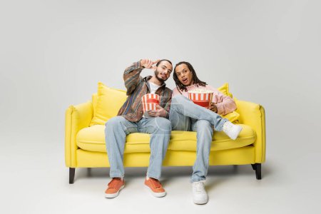 Vollbärtiger Mann, der mit dem Finger zeigt, während er einen Film mit einem multirassischen Freund mit Popcorn auf grauem Hintergrund ansieht