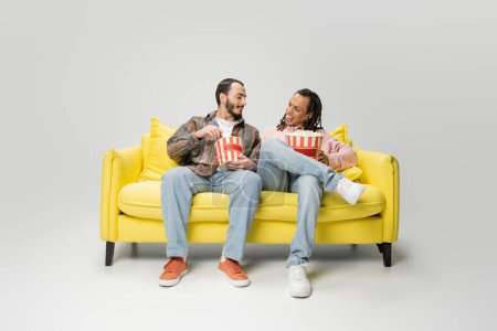 in voller Länge stilvolle multiethnische Männer, die einander anlächeln, während sie mit Eimern Popcorn auf einer gelben Couch auf grauem Hintergrund sitzen