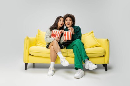 volle Länge der erstaunten interrassischen Frauen, die Popcorn in der Hand halten, während sie den Film auf der gelben Couch auf grauem Hintergrund ansehen