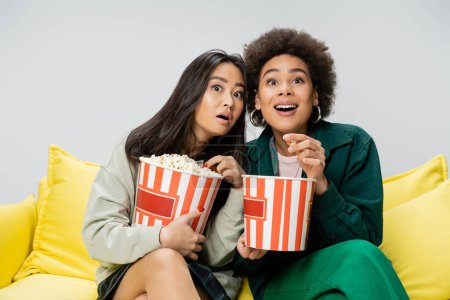 verblüffte interrassische Frauen, die Eimer Popcorn in der Hand halten, während sie einen Film auf der gelben Couch sehen, isoliert auf grau