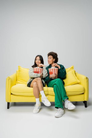 full length of cheerful multiethnic women eating popcorn and watching movie on yellow couch on grey background