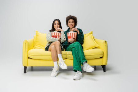 full length of smiling interracial friends eating popcorn and watching movie on yellow couch on grey background