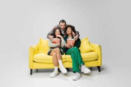 junger bärtiger Mann umarmt fröhliche interrassische Frauen, die mit Popcorn auf einer gelben Couch auf grauem Hintergrund sitzen