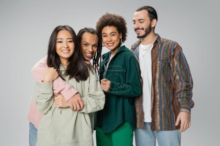 trendy multiracial man with dreadlocks embracing asian woman and smiling at camera near happy friends isolated on grey