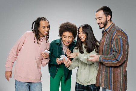 excited asian woman pointing at cellphone near smiling multicultural friends isolated on grey