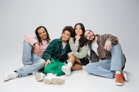 Téléchargez les photos : Full length of stylish multiethnic friends smiling at camera while sitting on grey background - en image libre de droit