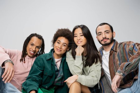 glückliche multiethnische Freunde in trendigen Klamotten lächeln in die Kamera, während sie isoliert auf grau sitzen