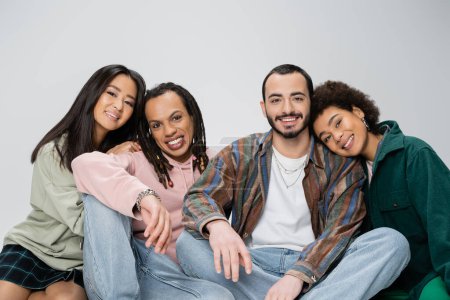 fröhliche multikulturelle Freunde im trendigen Outfit lächeln in die Kamera, während sie isoliert auf grau sitzen