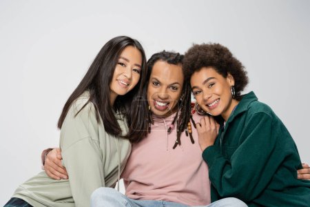 cheerful multiracial man with dreadlocks embracing asian and african american women isolated on grey