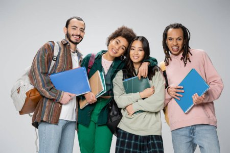 Photo for Happy multiethnic students with notebooks and backpacks smiling at camera isolated on grey - Royalty Free Image