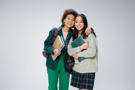 pretty interracial students with copybooks and backpacks smiling at camera isolated on grey
