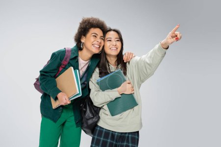 smiling asian student with copybooks looking away and pointing with finger near cheerful african american friend isolated on grey
