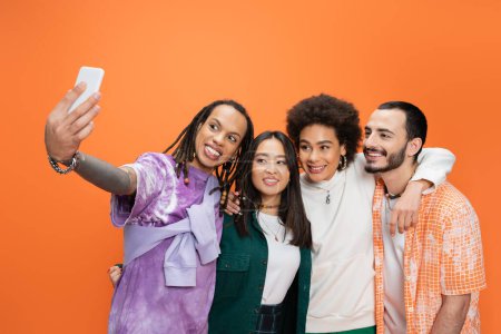 Téléchargez les photos : Trendy man with dreadlocks taking selfie with happy multicultural friends isolated on orange - en image libre de droit