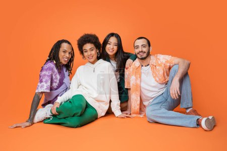 alegres amigos multiculturales en ropa elegante sentado y mirando a la cámara sobre fondo naranja